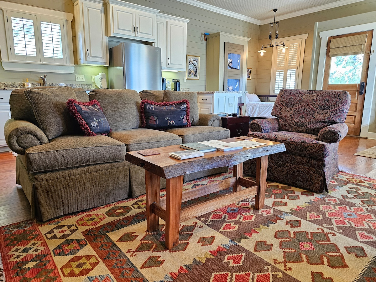 A cozy living area features a pair of plush sofas adorned with decorative pillows. A rustic wooden coffee table sits in the center, displaying a stack of magazines. The space is complemented by a colorful area rug and natural light filtering through the window.