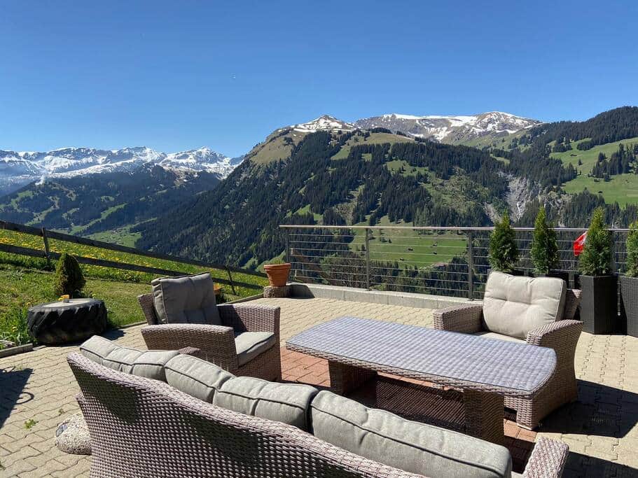 A spacious terrace is furnished with comfortable seating arrangements, including a light brown wicker set surrounding a rectangular coffee table. The backdrop features panoramic views of the vibrant alpine landscape, with green hills and snow-capped peaks under a clear blue sky.