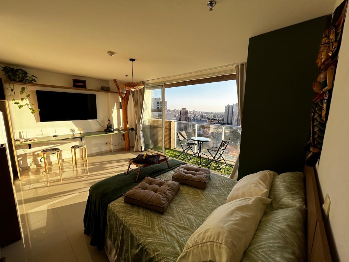 A spacious room features a large bed with soft linens and decorative pillows. A modern entertainment unit is situated nearby, and a small dining table is visible. Floor-to-ceiling windows provide a view of the balcony, where outdoor seating is arranged.