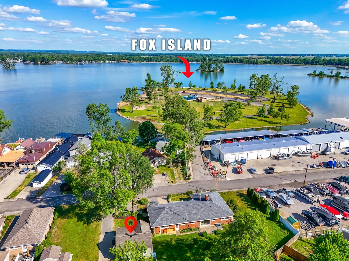 Aerial view captures Fox Island, surrounded by the calm waters of Indian Lake. Nearby cottages and the expansive shoreline are visible, with green trees dotting the landscape. The vibrant blue sky adds to the scenic backdrop of this tranquil area, highlighting the proximity to nature.