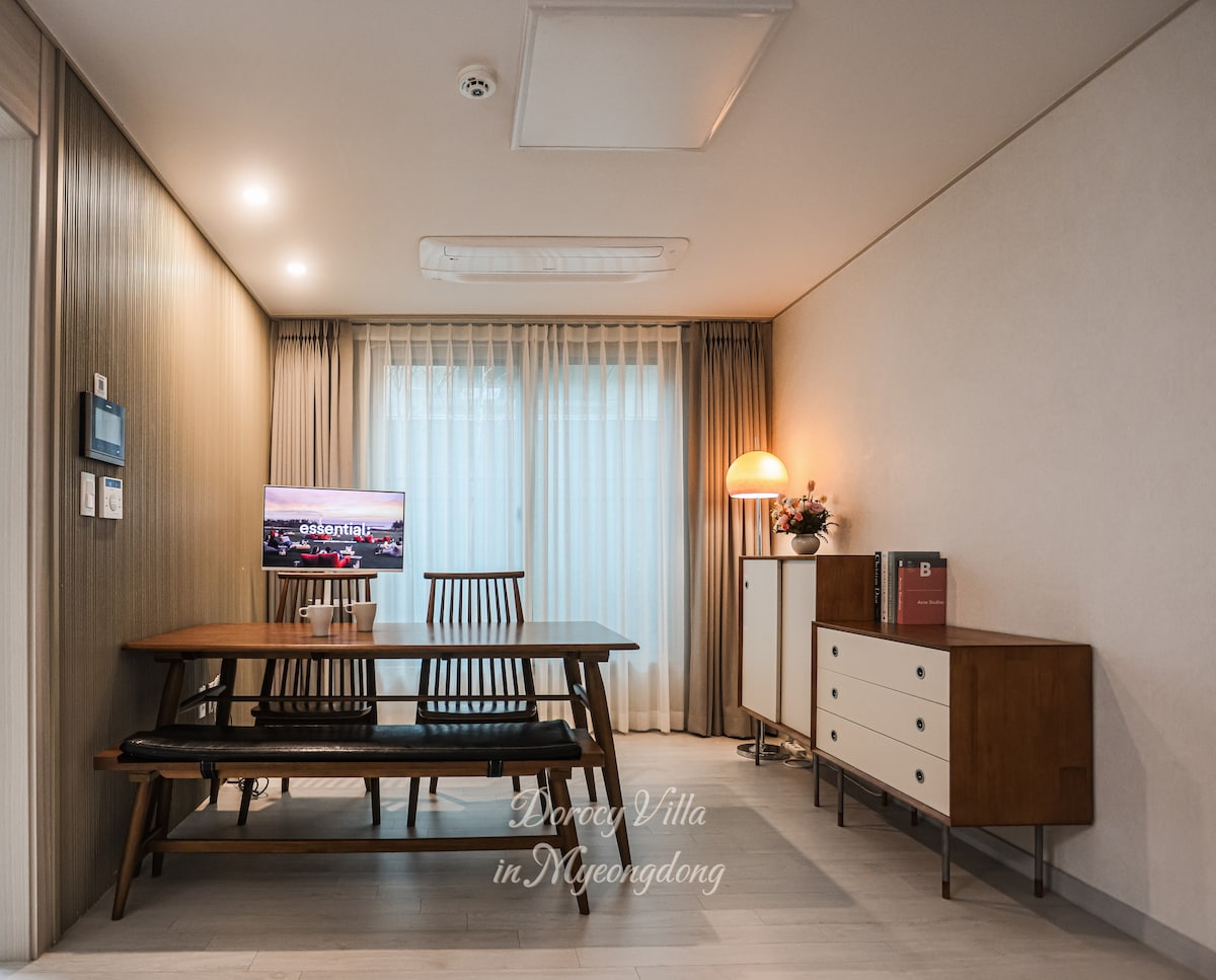 A bright living area is furnished with a wooden dining table and matching chairs, providing a comfortable space for meals. A television is positioned against the wall, while a sleek sideboard adds storage. Natural light floods the room through sheer curtains.
