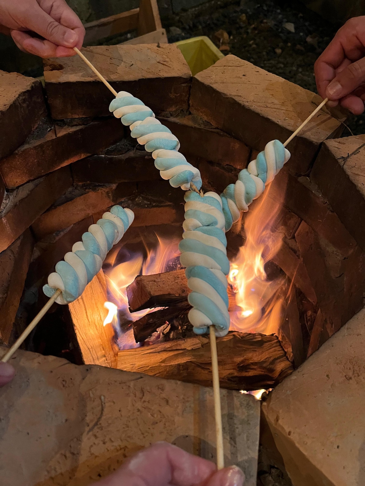 Multiple skewers with twisted marshmallows are held above a warm, glowing fire within a circular stone fire pit. Flames dance among the wooden logs, creating a cozy atmosphere for gathering and enjoying treats.