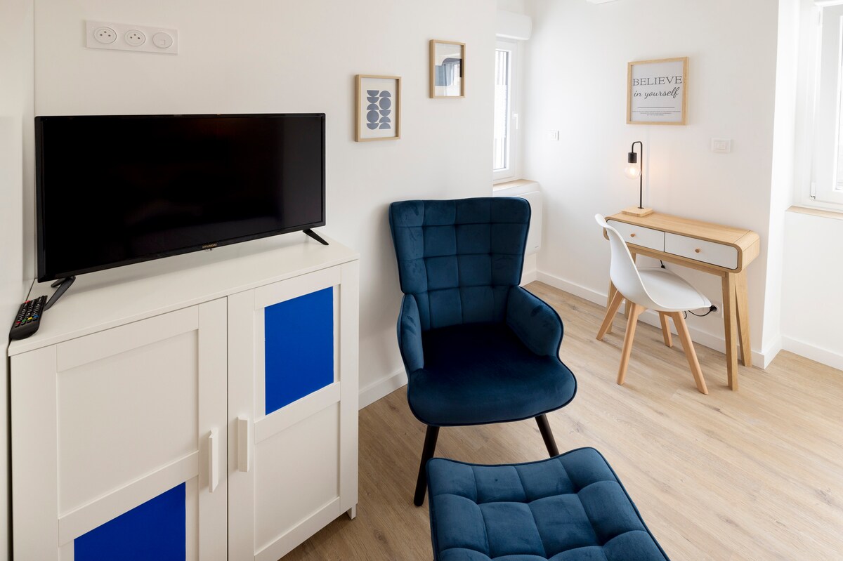 A bright living space features a wall-mounted television and a modern desk with a stylish lamp. A comfortable blue chair, paired with a matching ottoman, invites relaxation. Natural light enters through the windows, enhancing the clean and contemporary design of the room.