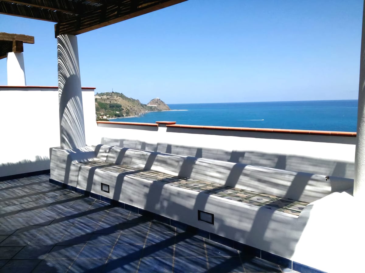 A spacious terrace features a long, built-in seating area with soft cushions, providing a comfortable spot for relaxation. The ocean stretches out in the background, showcasing vibrant blue waters and a clear sky. Sunlight creates a pattern of shadows on the tiled flooring.