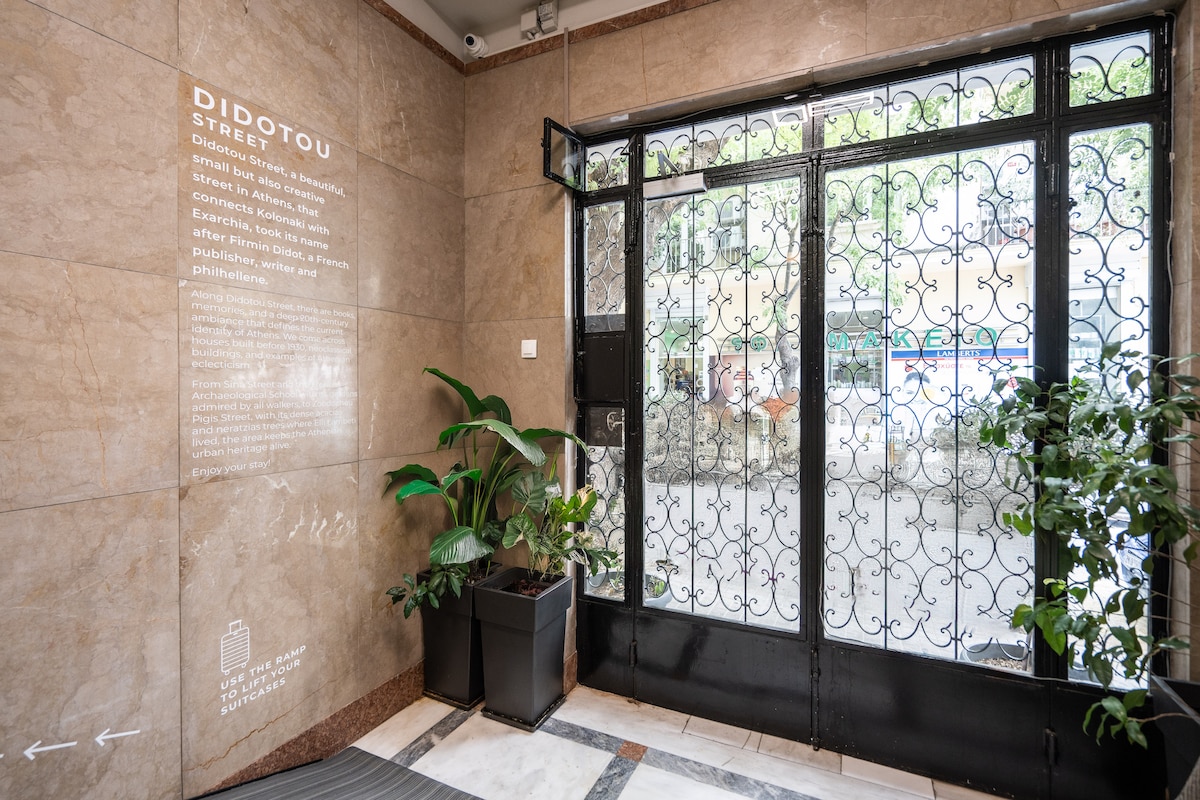 The entrance area showcases an intricate wrought iron door with clear glass panels, allowing natural light to enter. Potted greenery adds a refreshing touch to the space, complementing the warm-toned marble walls and tiled floor. Informative signage is displayed on the wall beside the door.