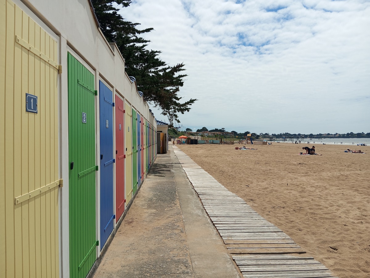 A sandy beach is lined with colorful cabin doors in a variety of hues, including green, yellow, and red. A wooden walkway runs alongside the beach, leading to areas where individuals are placed on the sand, enjoying leisure activities under a partly cloudy sky.