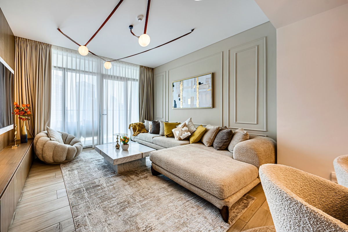 The living area features a spacious sectional sofa adorned with various textured cushions, offering comfort. A minimalist coffee table is centered atop a soft area rug. Large windows allow natural light to fill the space, while light curtains enhance the inviting atmosphere.