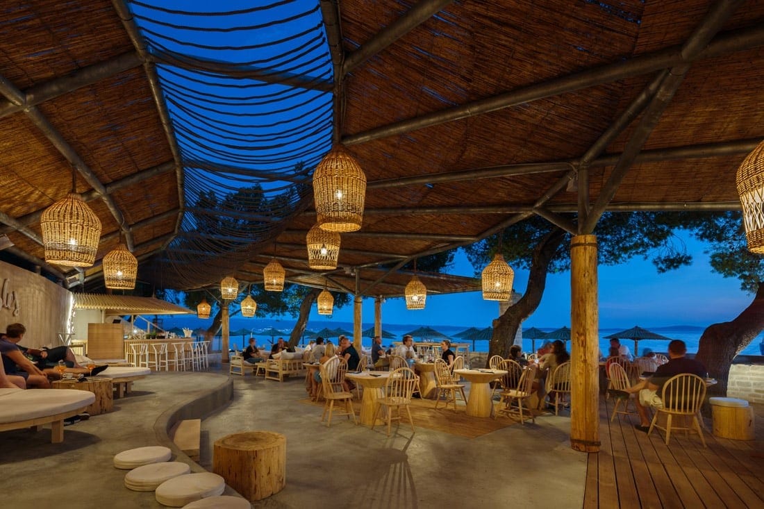 An outdoor dining space features a thatched roof supported by wooden beams, illuminated by hanging lanterns. Guests are seated at various tables, with a calming view of the sea in the background. Soft seating areas invite relaxation under the evening sky.