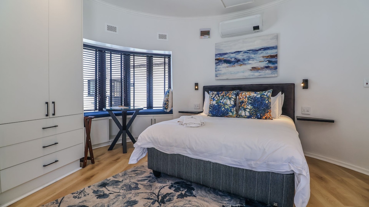 A spacious sleeping area featuring a large bed with decorative pillows. Natural light enters through a curved bay window with horizontal blinds. A small round table and a wooden chair are present, alongside a minimalistic wall-mounted shelf, enhancing the room's layout.