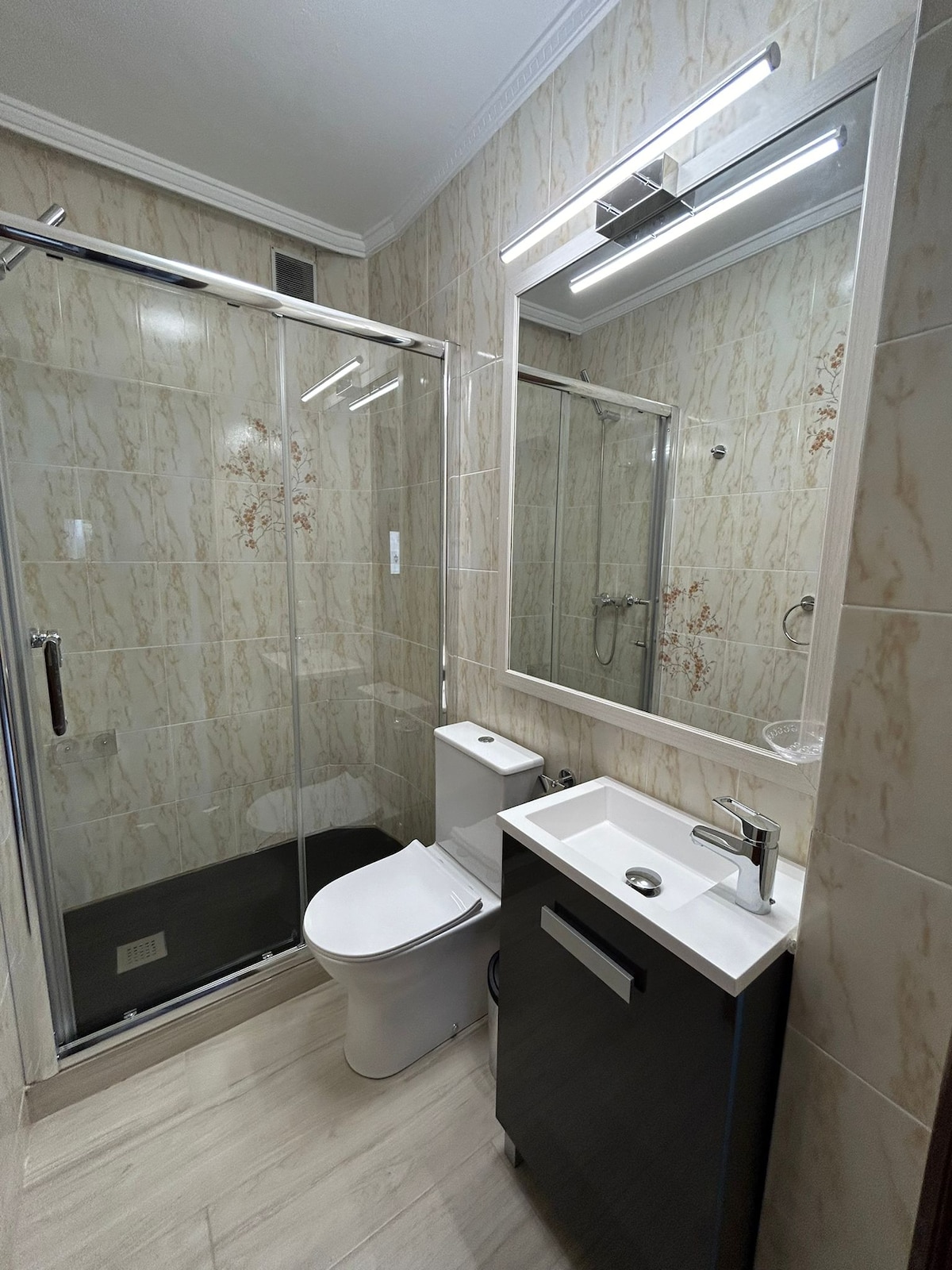 The bathroom features a glass-enclosed shower and a modern toilet. A wall-mounted mirror is positioned above a compact sink with a sleek faucet. Light-colored tiled walls and floor contribute to a clean and bright environment.