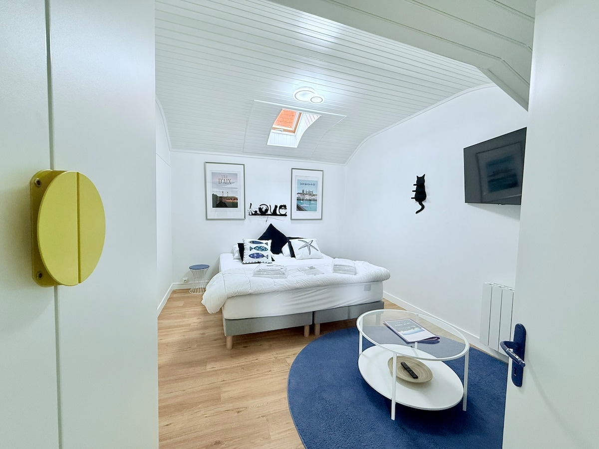 A bright bedroom features a spacious bed with white bedding and decorative pillows. Artwork adorned walls provide aesthetic appeal, while a television is mounted for entertainment. A round coffee table sits on a blue rug, creating a cozy gathering space. Natural light enters through a skylight.