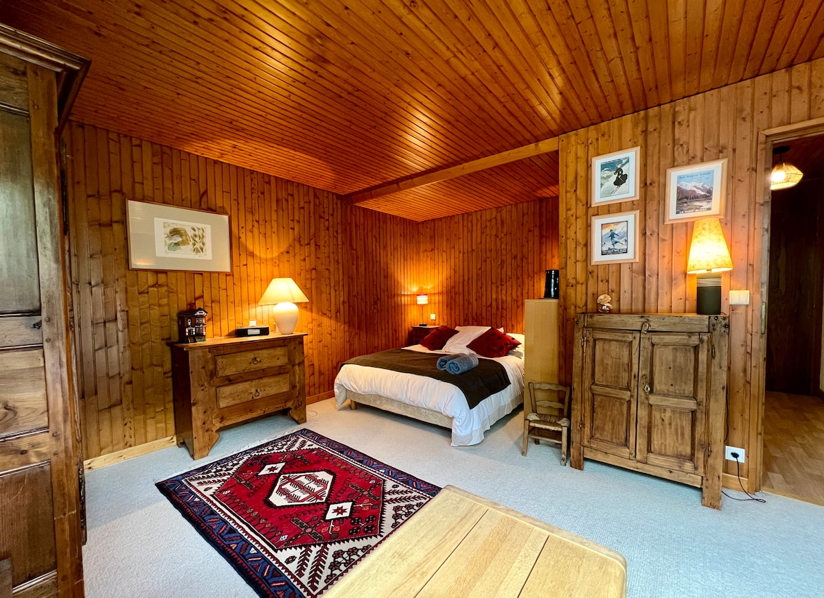 The spacious room features wooden paneling throughout, creating a warm atmosphere. A comfortable bed is positioned against the far wall, accompanied by bedside lamps. A decorative rug complements the space, with wooden furniture providing storage. Natural light enters from an adjacent doorway.