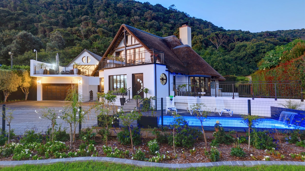 A charming cottage with a thatched roof is surrounded by lush greenery. The property features an outdoor pool and is set within a well-maintained garden. A spacious driveway leads to an attached garage, while outdoor seating options invite relaxation.
