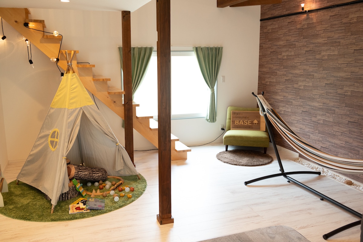 A cozy play area is equipped with a children's tent decorated in yellow and gray, surrounded by colorful toys on a green area rug. A comfortable hammock is positioned nearby, while natural light enters through windows adorned with light green curtains.