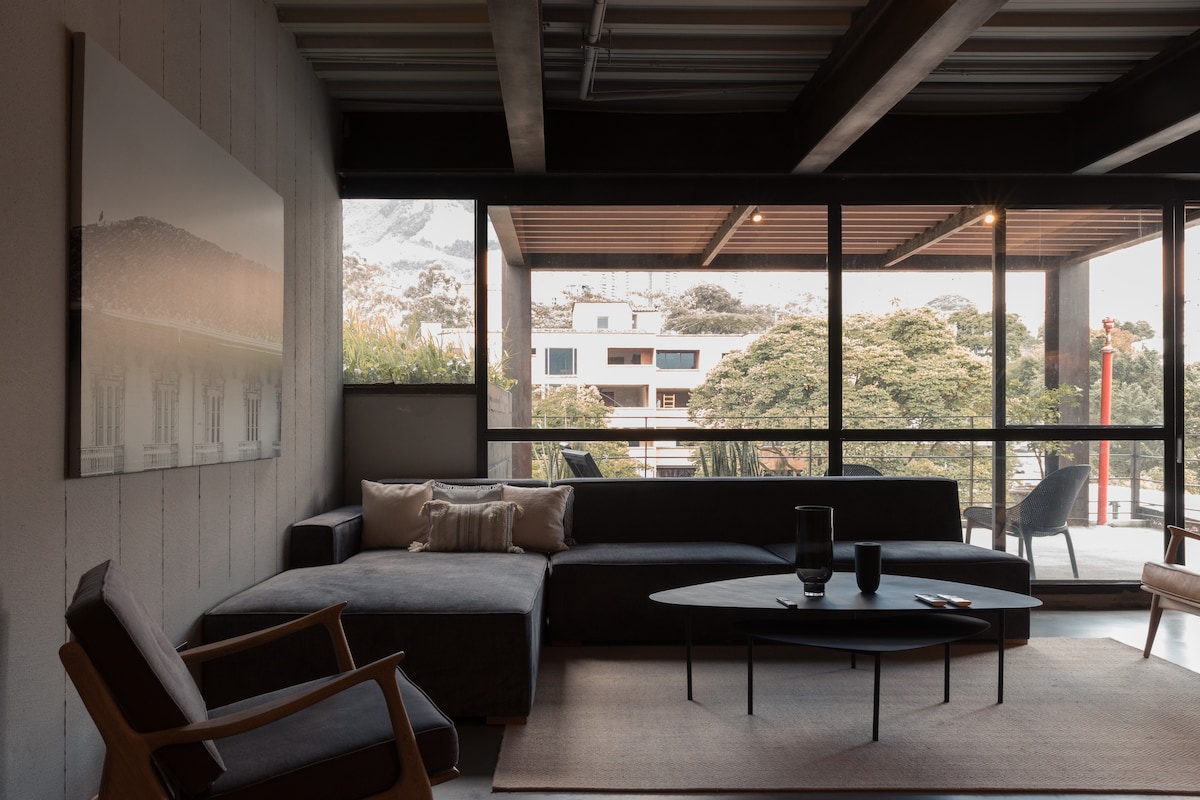 A bright living area is presented with large windows, offering views of the surrounding landscape. An L-shaped sectional sofa is complemented by a circular coffee table. Two chairs are positioned nearby, creating a comfortable space for relaxation and conversation.