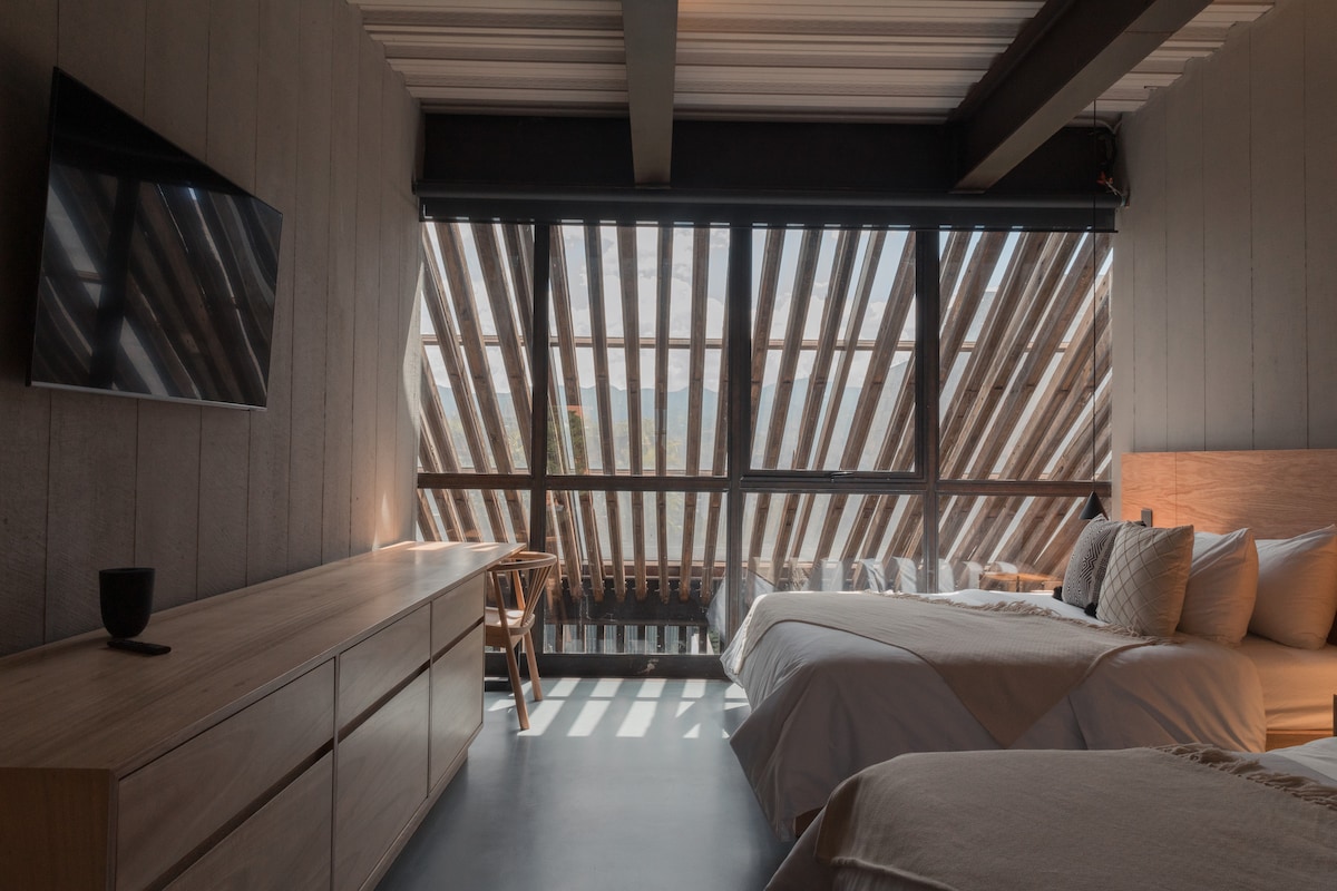 A spacious room is arranged with two beds positioned beside a large window featuring distinctive wooden slats. Soft natural light filters through, casting lines and patterns on the floor. A mounted television and a wooden cabinet are visible, contributing to the room's minimalist design.