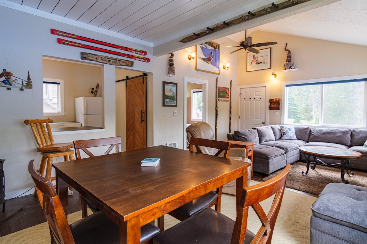 A dining area is furnished with a wooden table surrounded by four chairs, creating a communal space. The living room features a spacious gray sectional sofa, accentuated by natural light from a large window. Scenic artwork and decorative skiing equipment adorn the walls.