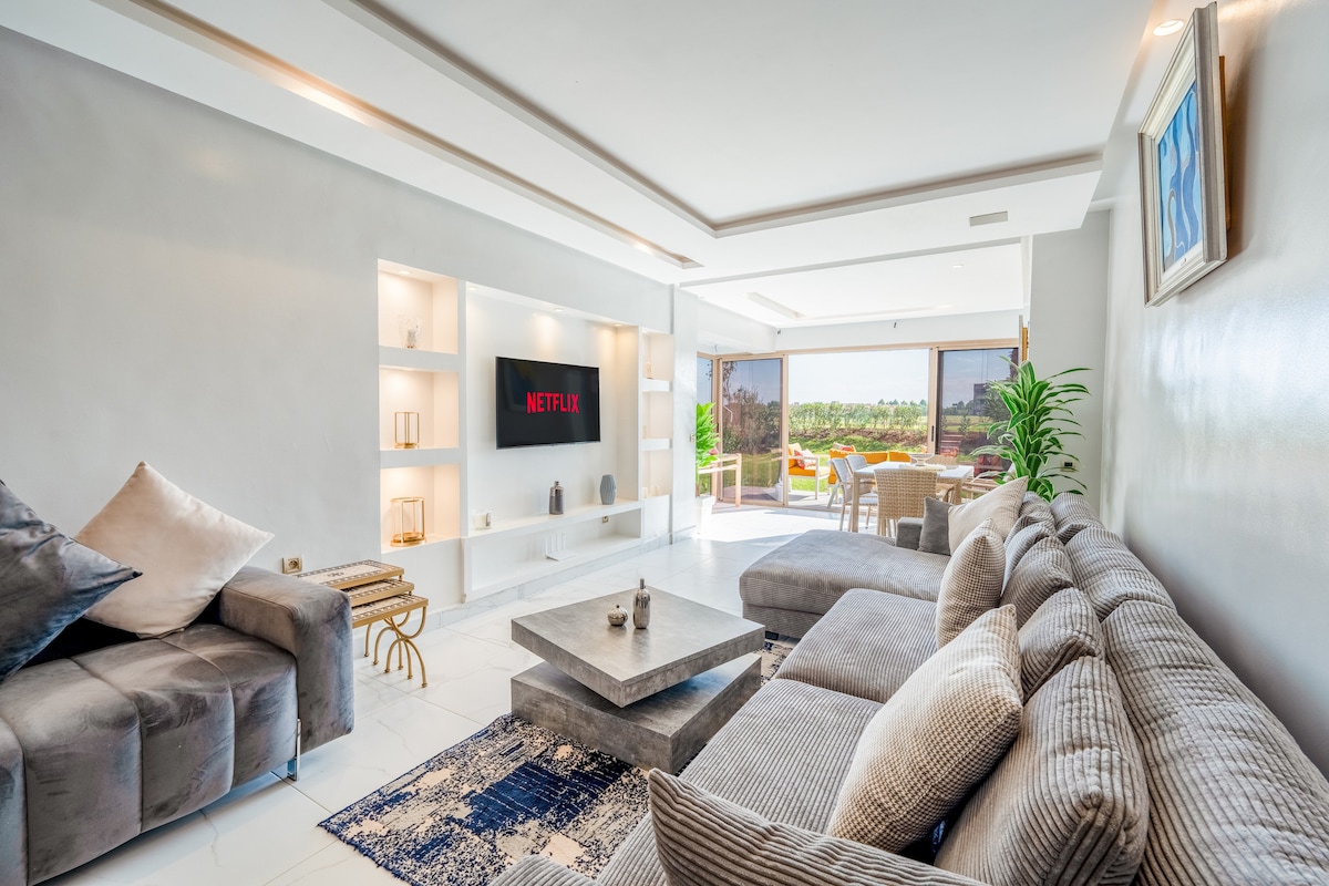 A stylish living area features a comfortable sectional sofa and a modern coffee table. A flat-screen TV mounted on the wall displays a streaming service. Large glass doors lead to a terrace with outdoor seating, allowing natural light to fill the space.