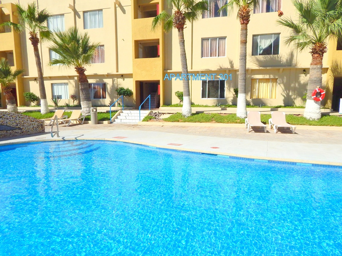 The image displays a clear blue pool in the foreground, surrounded by a well-maintained patio and palm trees. In the background, a multi-story building with several windows is shown, indicating Apartment 301. Lounge chairs are positioned beside the pool, inviting relaxation.
