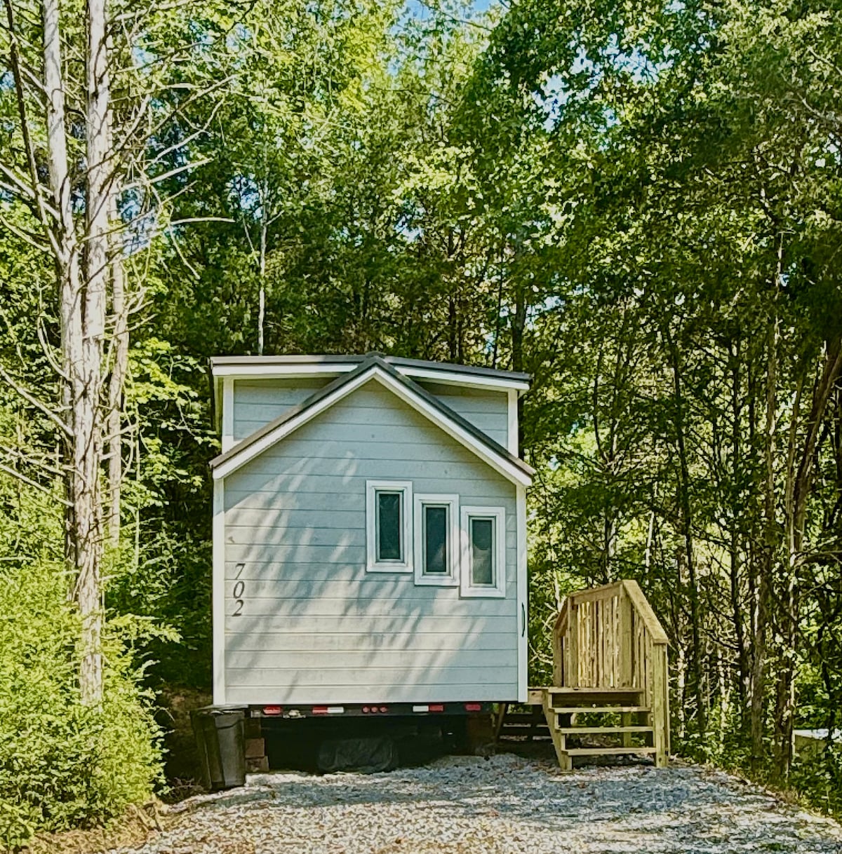 The exterior of a tiny home cabin is visible, featuring a light-colored facade and a sloped roof. Large windows are positioned at the front, allowing natural light to fill the interior. A wooden staircase leads up to the entrance, surrounded by greenery.