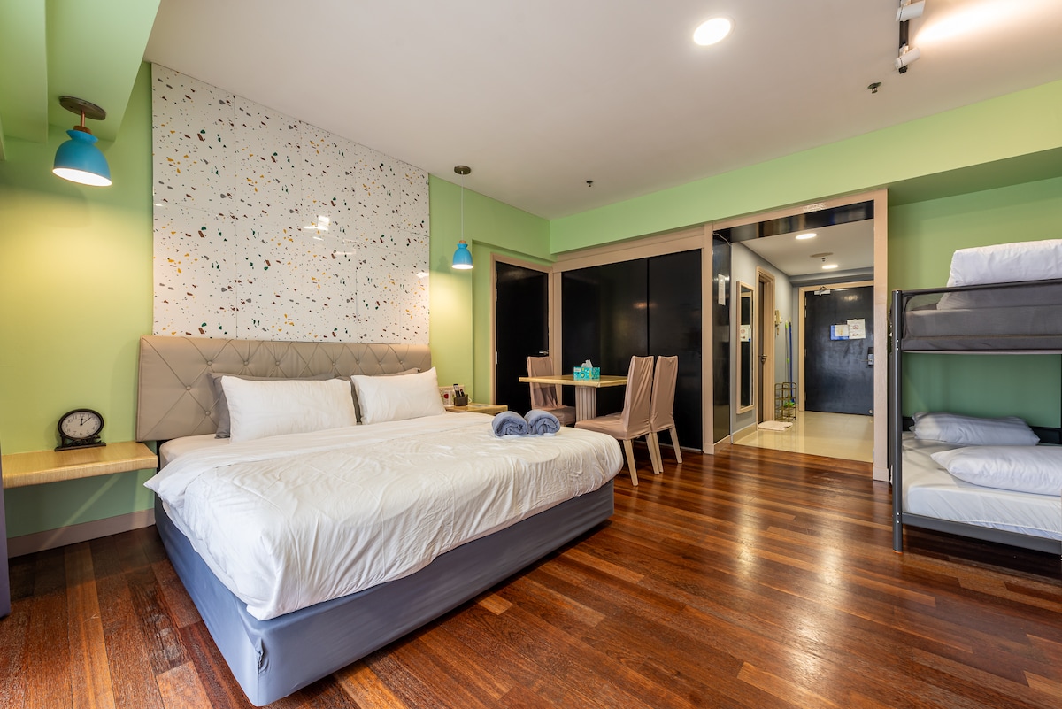 A studio suite features a spacious bed with a light-colored headboard and soft linens. A cozy bunk bed is positioned nearby. A dining table with four chairs is present, and the walls display a modern speckled design. Warm wooden floors enhance the overall ambiance.