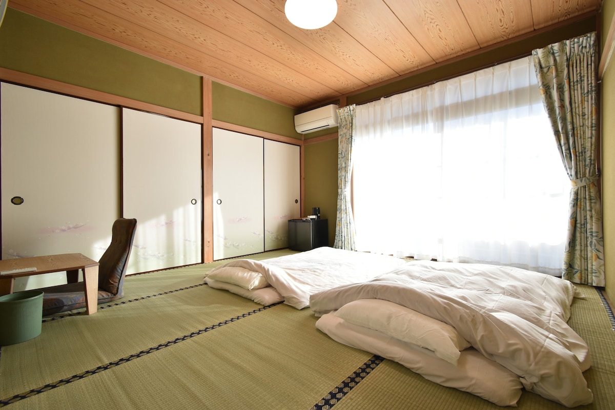 A spacious traditional Japanese-style room features tatami flooring with two laid-out futons. A simple wooden table accompanies a comfortable chair. Light streams through large windows adorned with curtains, creating a serene atmosphere, while air conditioning ensures a comfortable environment.