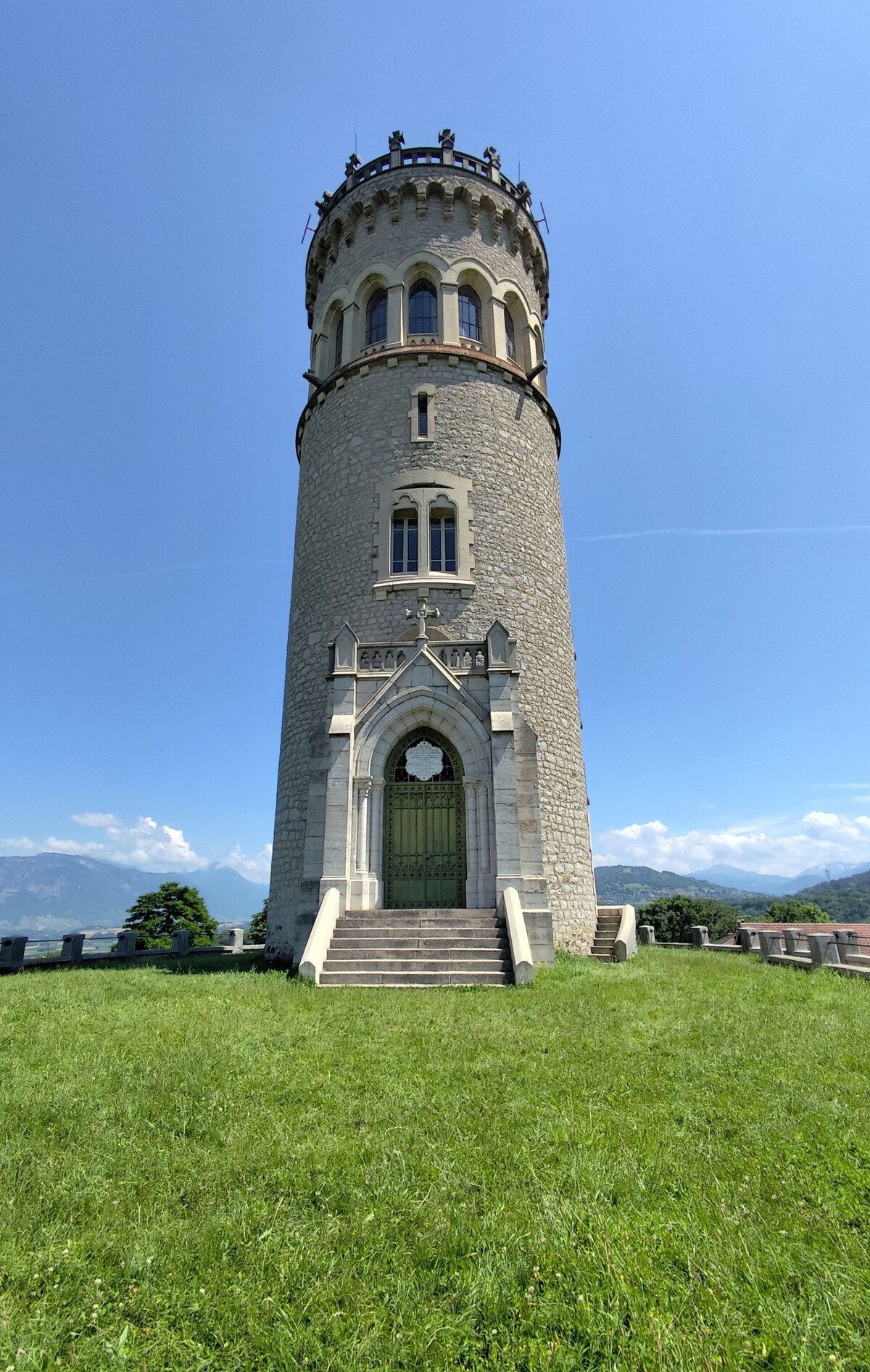 A stone tower stands majestically against a clear blue sky, featuring intricate architectural details. The tower is framed by lush greenery and offers panoramic views of surrounding mountain ranges. A pathway leads to the entrance, highlighting the structure's historical significance.