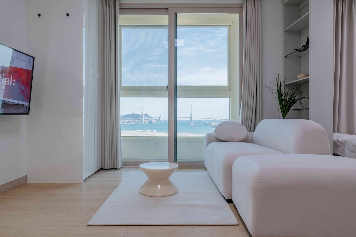 A modern living space features a white sectional sofa and a circular coffee table. Large windows provide a panoramic view of the ocean and nearby bridge, while sheer curtains allow natural light to brighten the room.