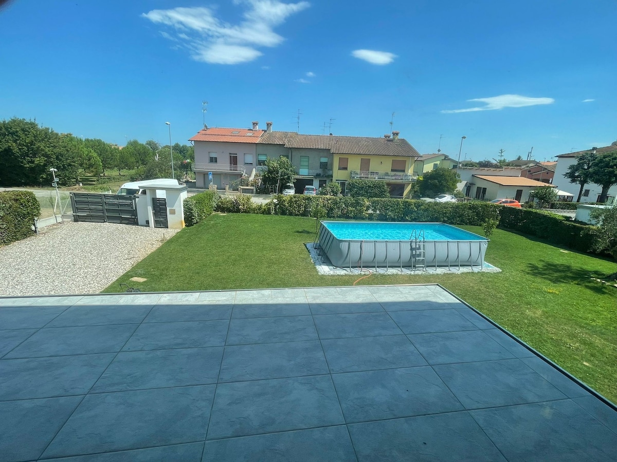 A spacious outdoor area is visible, featuring a well-maintained grass lawn and a rectangular above-ground pool. Surrounding greenery, including hedges and trees, provides a natural backdrop, alongside a gravel driveway leading to the entrance of the property.