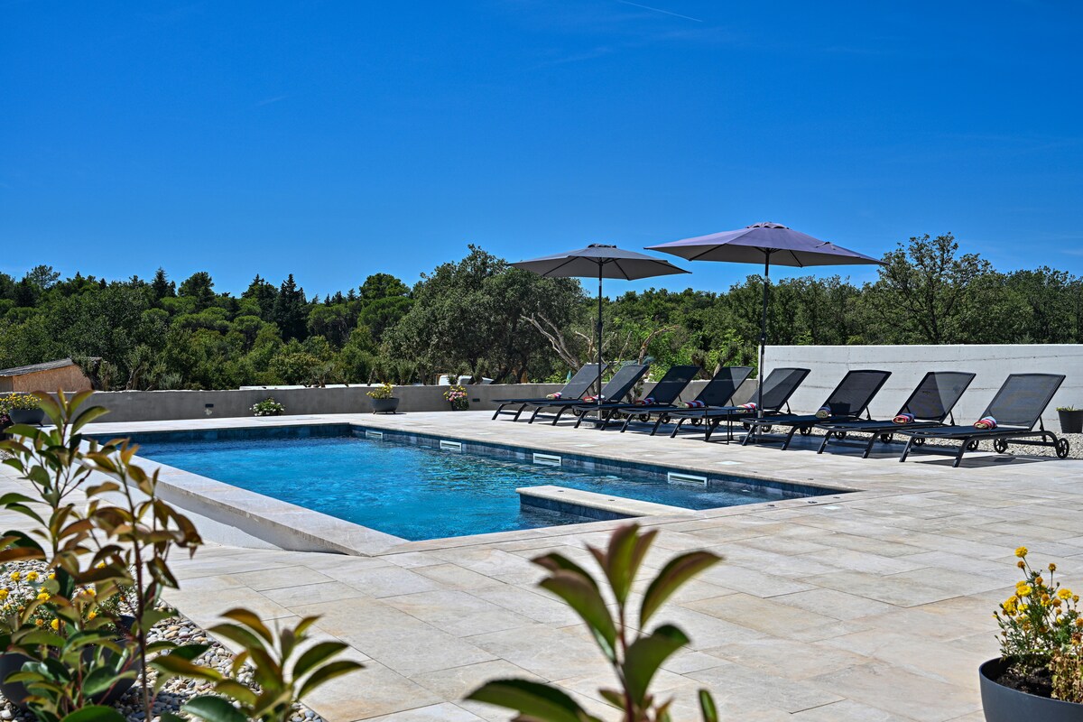 A swimming pool is set within a spacious stone patio, surrounded by sun loungers. Two umbrellas provide shade, while greenery lines the background, creating a peaceful outdoor setting. The clear blue sky complements the tranquil atmosphere.
