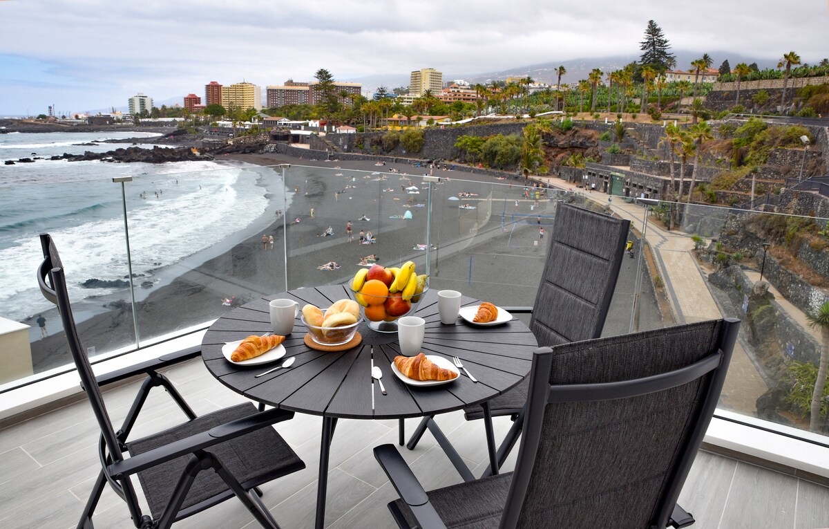 A terrace table is set with breakfast items, including pastries and fruit, overlooking a beach with black sand. The scene features a view of the ocean and a shoreline with people enjoying the beach. The surrounding area is lined with palm trees and buildings.