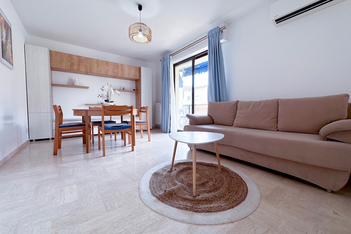 A spacious living area features a comfortable sofa and a round coffee table atop a woven rug. A wooden dining table is surrounded by chairs, complemented by shelves holding decorative items. Natural light filters through large windows, and light-colored curtains enhance the openness.