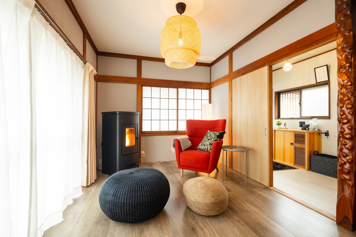 A traditional Japanese living room is displayed, featuring a stylish red armchair and two poufs in neutral tones. Natural light enters through a grid-patterned window, illuminating the space. A sleek pellet stove offers warmth, while wooden accents and soft textures create a relaxing environment.
