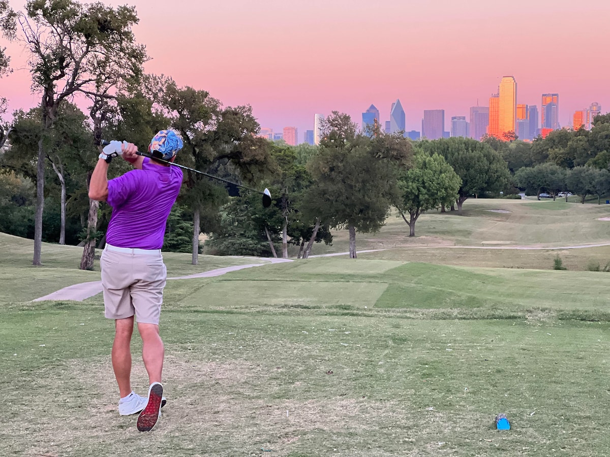 A golfer is captured mid-swing on a lush green golf course, with the Dallas skyline visible in the background. Soft hues of pink and orange illuminate the sky at sunset, contrasting with the vibrant greenery of the course.