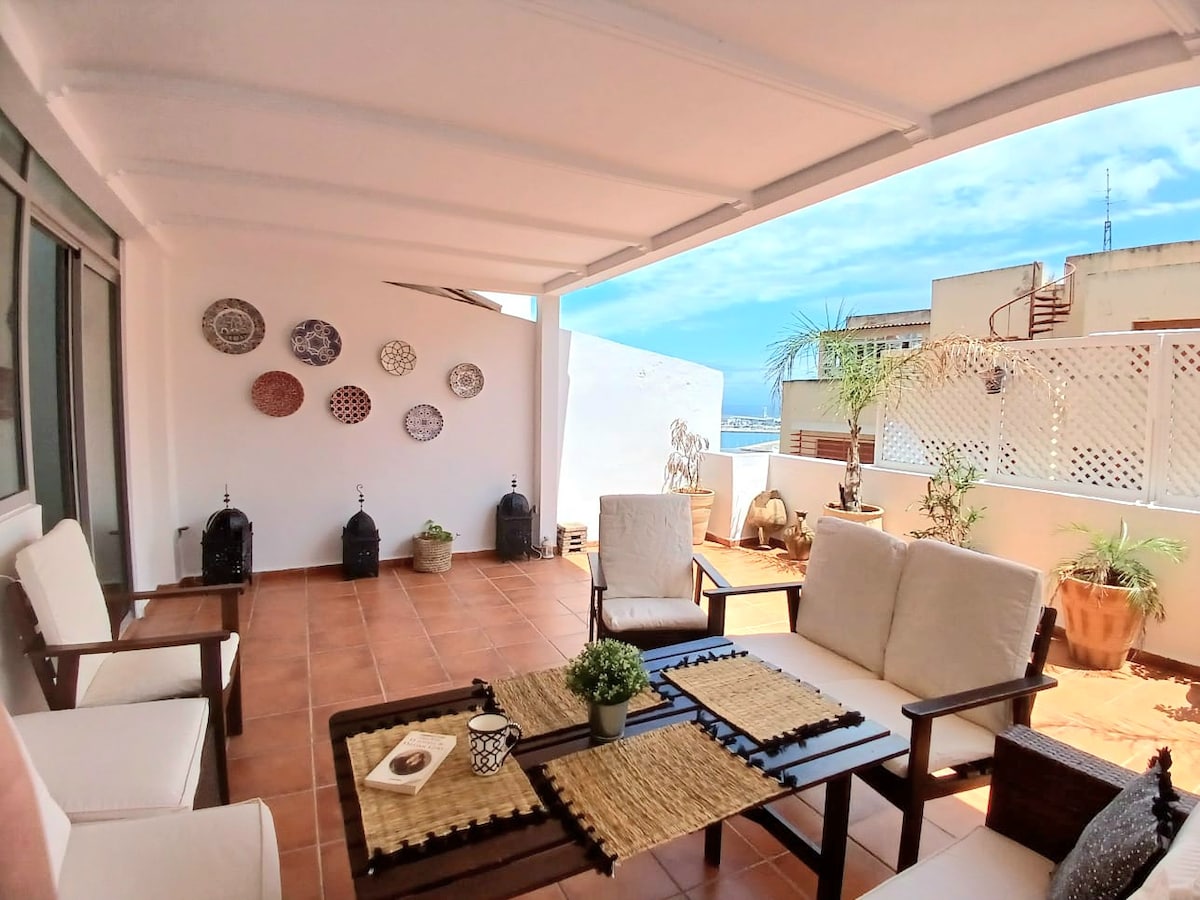 A spacious terrace is visible, furnished with comfortable seating options and a wooden table. Decorative plates adorn the wall, complemented by potted plants. The sea can be seen in the distance, and a clear blue sky adds to the relaxed atmosphere.