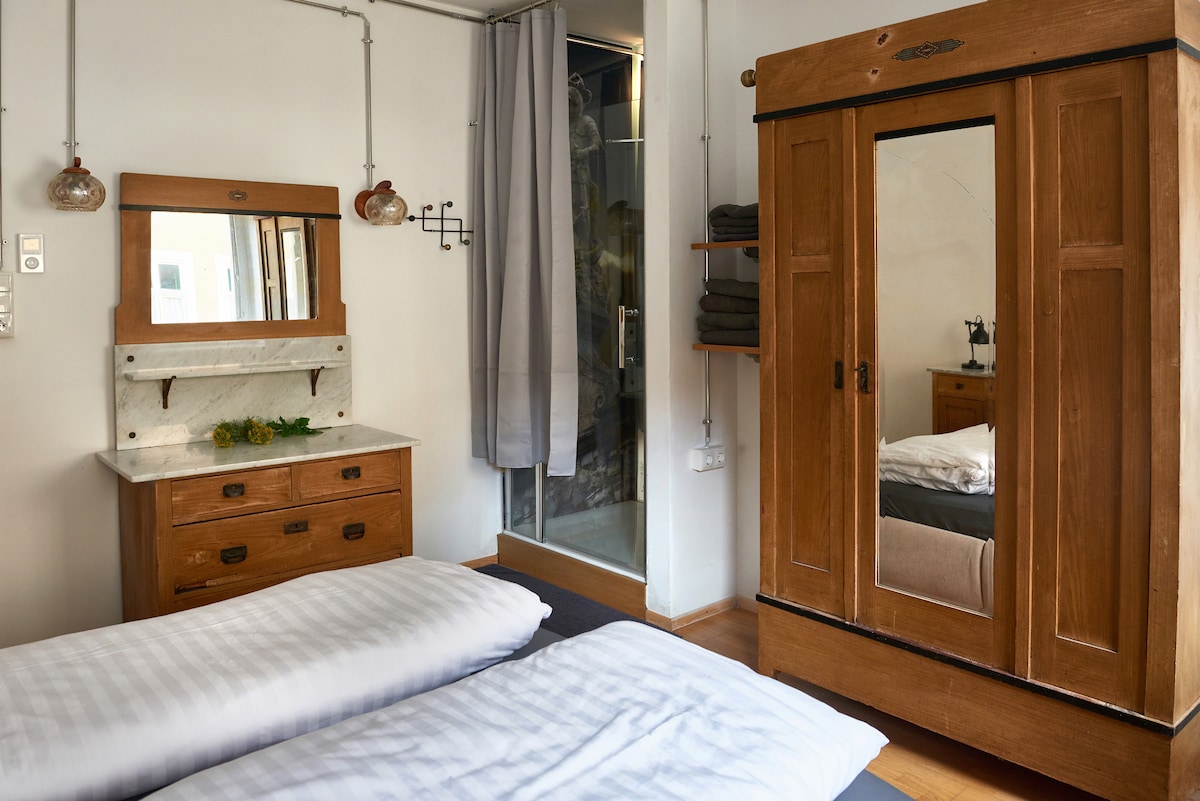 A cozy bedroom features two spacious beds with neatly arranged white linens. A vintage wooden wardrobe stands against the wall, displaying a mirrored door. A marble-topped dresser is accompanied by decorative wall-mounted lamps, while a curtain separates the room from a nearby bathroom.