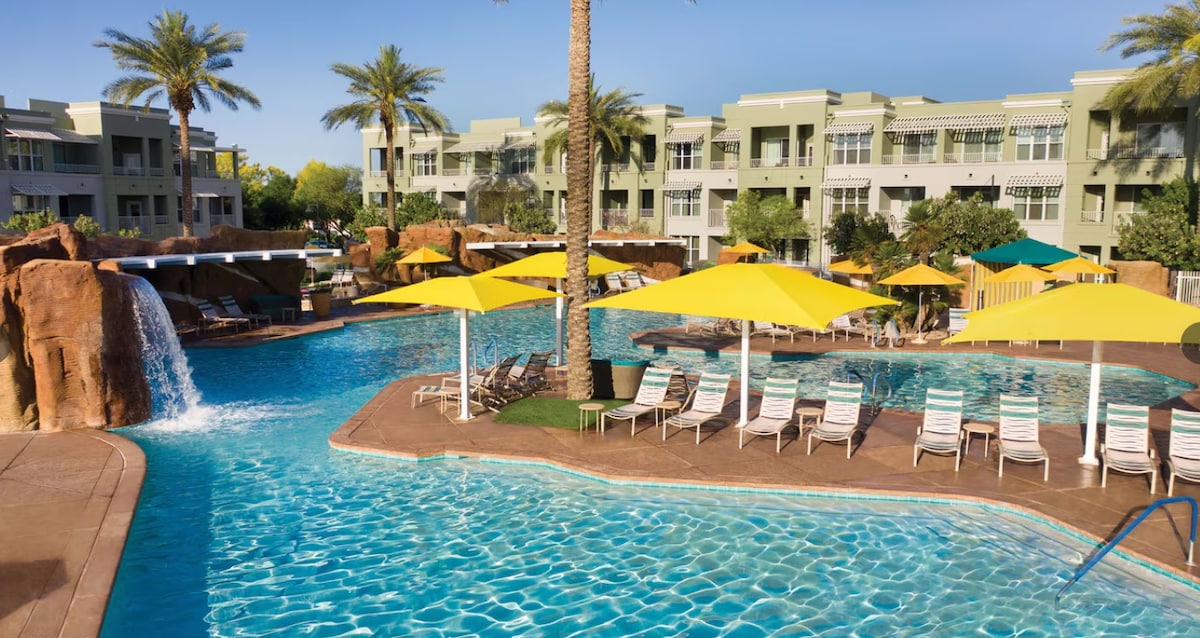 A large outdoor pool area is surrounded by palm trees and features a waterfall cascading into the water. Yellow umbrellas provide shade over several lounge chairs, while additional structures and greenery enhance the relaxing ambiance of the resort.