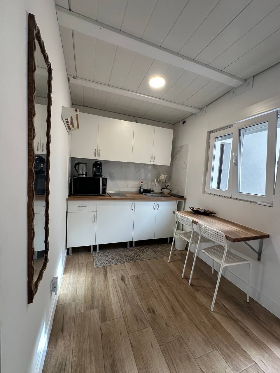 A bright and functional kitchen space features white cabinetry and a wooden countertop. Essential appliances include a coffee machine and microwave. A small wooden table with two chairs invites casual dining, while natural light streams in through two small windows.