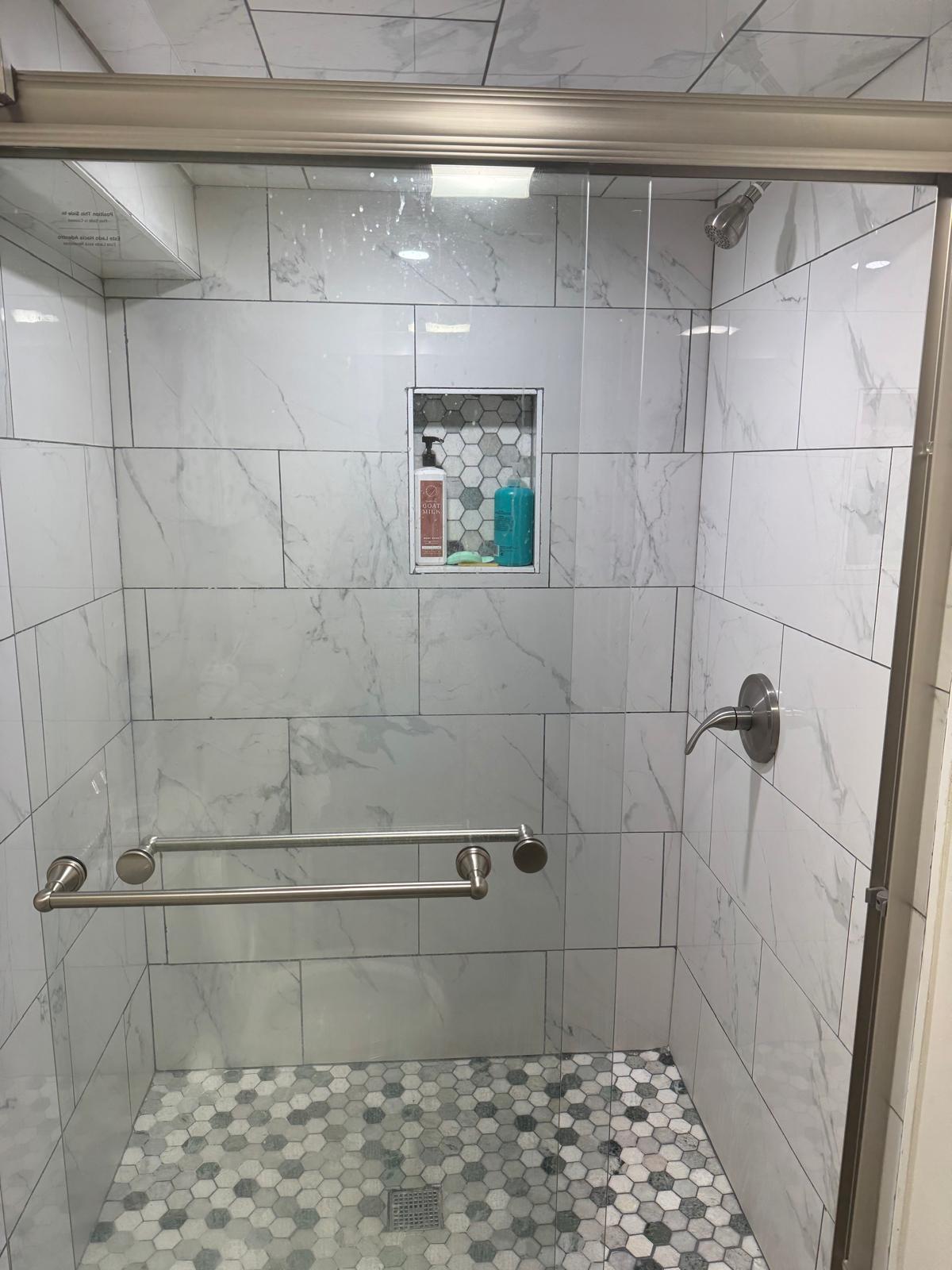 A modern shower area is enclosed by clear glass, featuring elegant white marble tiles. A built-in shelf holds personal care items. The floor is decorated with hexagonal tiles, and grab bars are positioned for safety and accessibility.