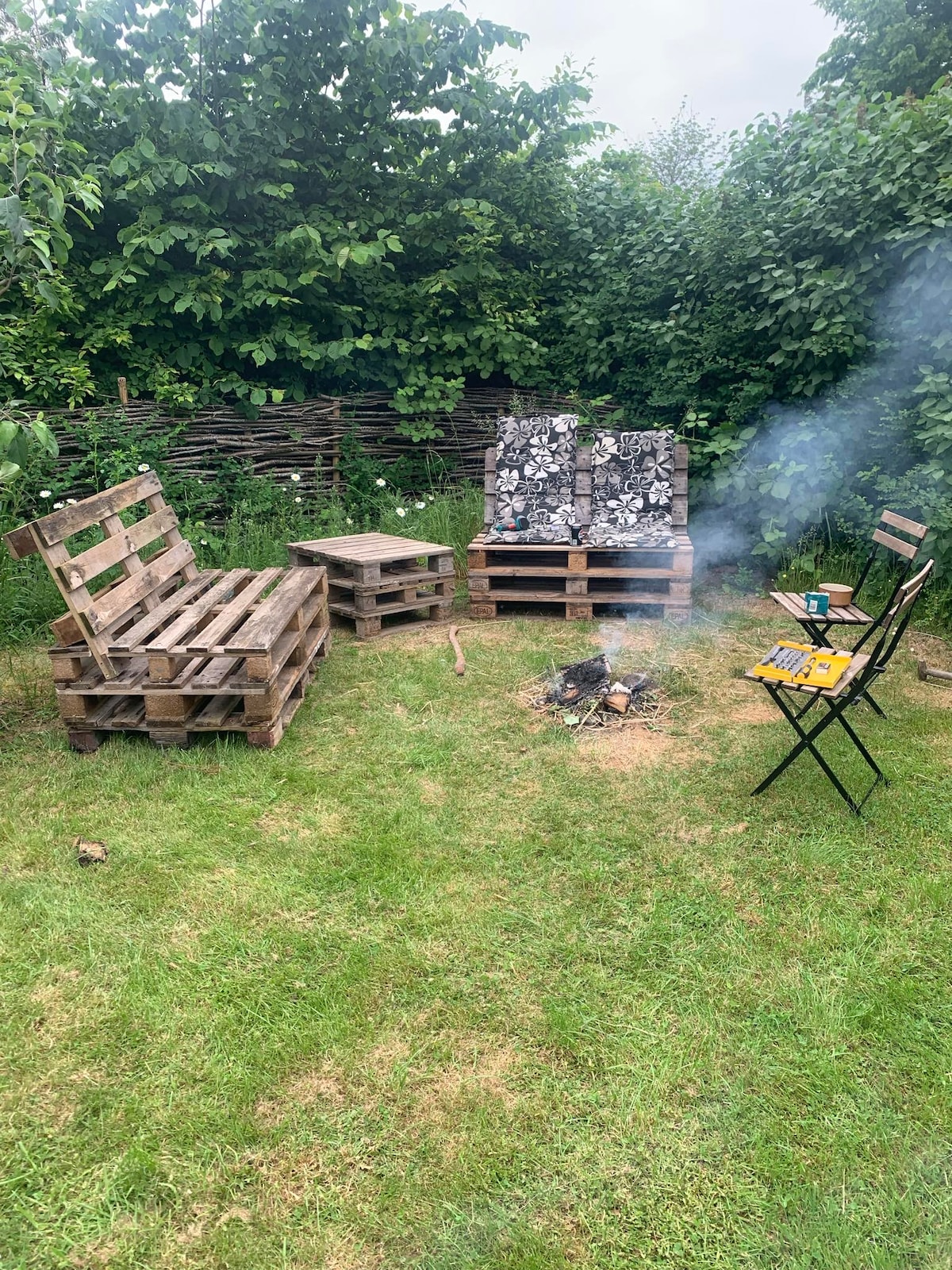 An outdoor seating area features furniture crafted from wooden pallets, including a coffee table and several low couches. A small campfire surrounded by a grassy area adds a sense of relaxation, with smoke gently rising amidst greenery in the background.