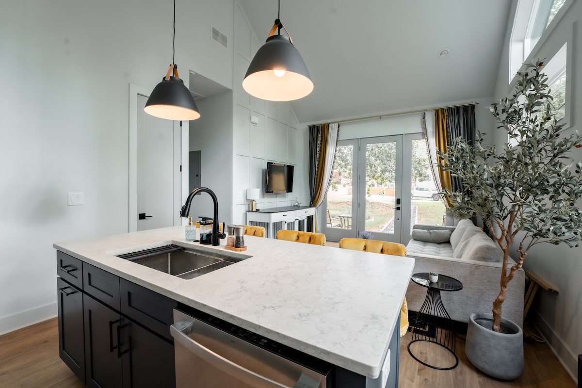 A modern kitchen island stands in the foreground, featuring a white marble countertop with a sink. Above, two pendant lights illuminate the space. The airy living area is visible in the background, furnished with a light-colored couch, a TV, and large windows revealing outdoor views.