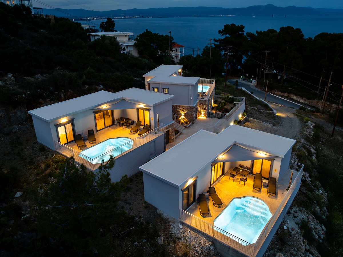 Three modern villas are shown, illuminated softly against the evening backdrop. Each villa is equipped with a private pool, surrounded by lounge chairs. The tranquil setting is complemented by the nearby coastline and distant sea view.