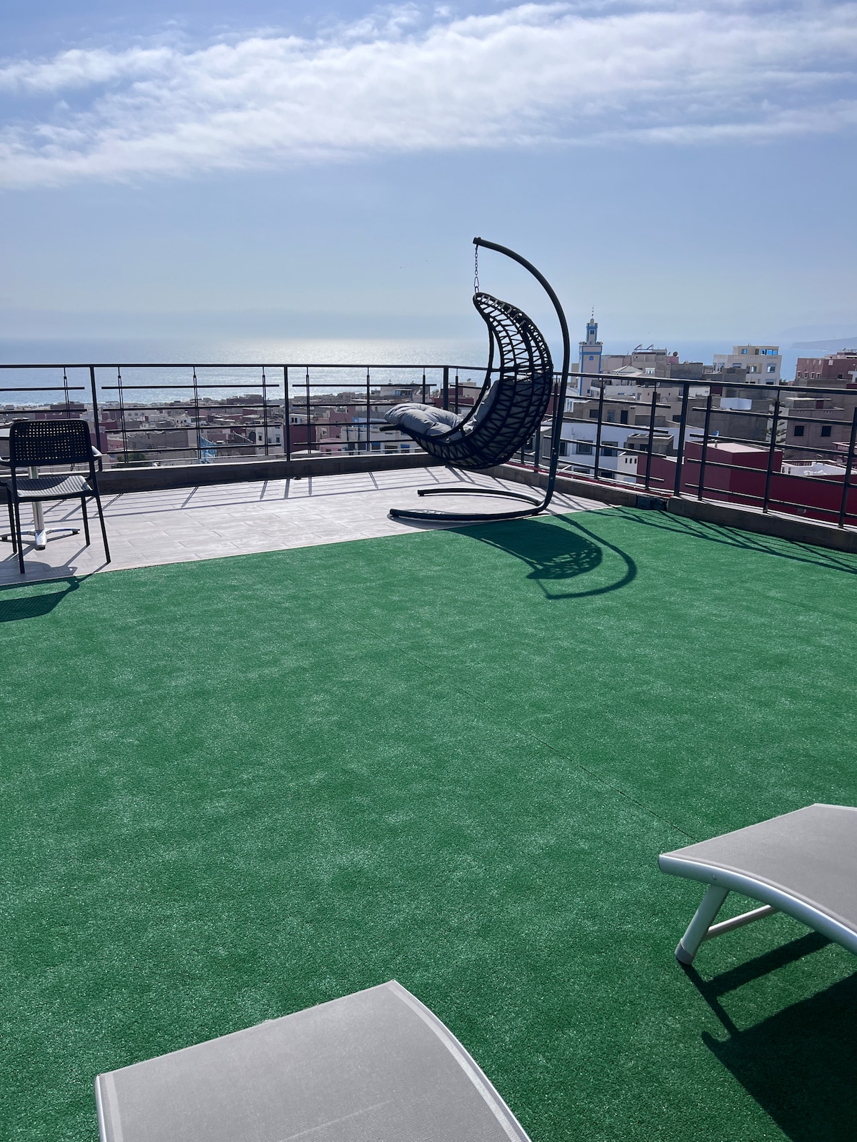 The rooftop terrace features artificial grass flooring, providing a comfortable area for relaxation. A suspended swing chair overlooks picturesque views of the sea and surrounding landscape. Tables and lounge chairs are arranged to accommodate guests, inviting enjoyment of the tranquil outdoor space.