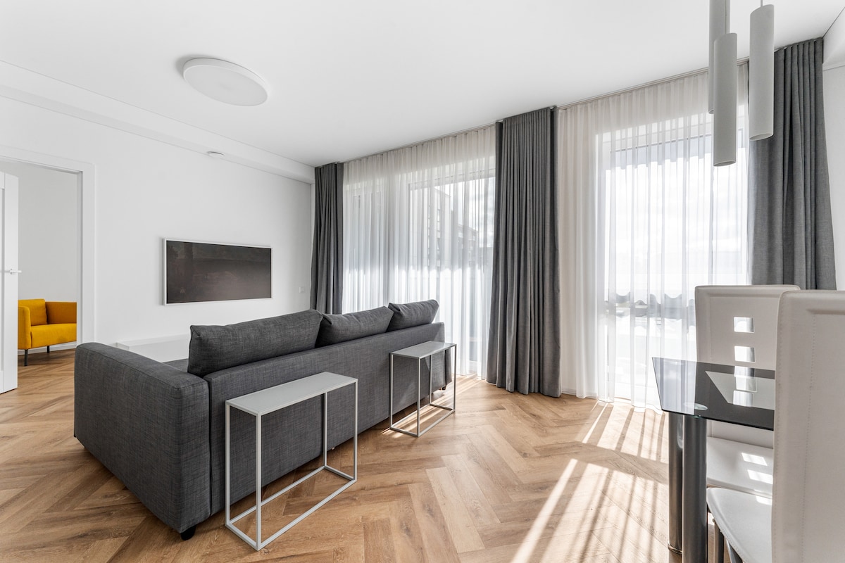 The living area features a gray sofa positioned in front of large windows with sheer curtains that allow natural light to filter in. A sleek side table stands beside the sofa, and a modern dining table with white chairs is visible in the background. Light wood flooring adds warmth to the space.