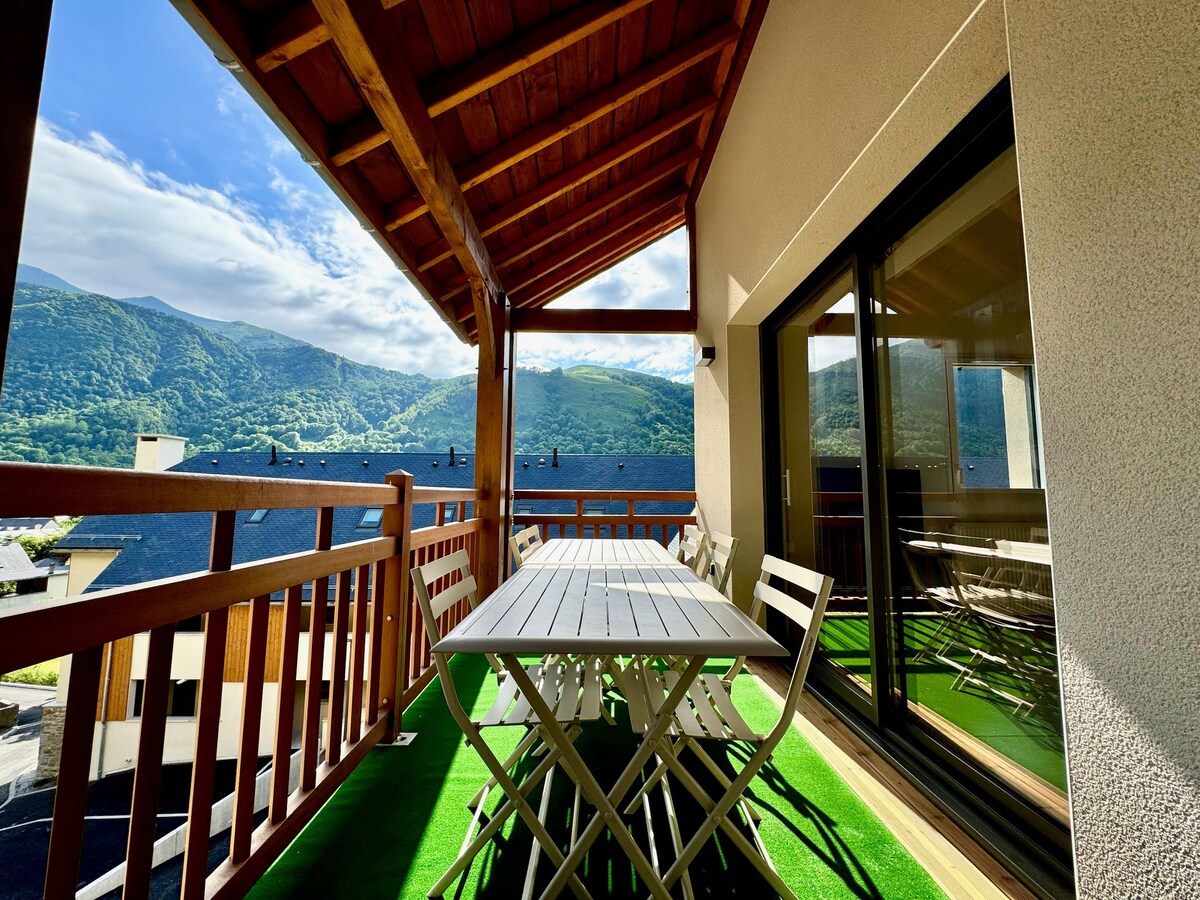 A balcony with a wooden overhead structure features a large table surrounded by chairs. The backdrop showcases expansive mountain views under a clear blue sky. Sunlight illuminates the area, creating a pleasant outdoor space for relaxation and dining.