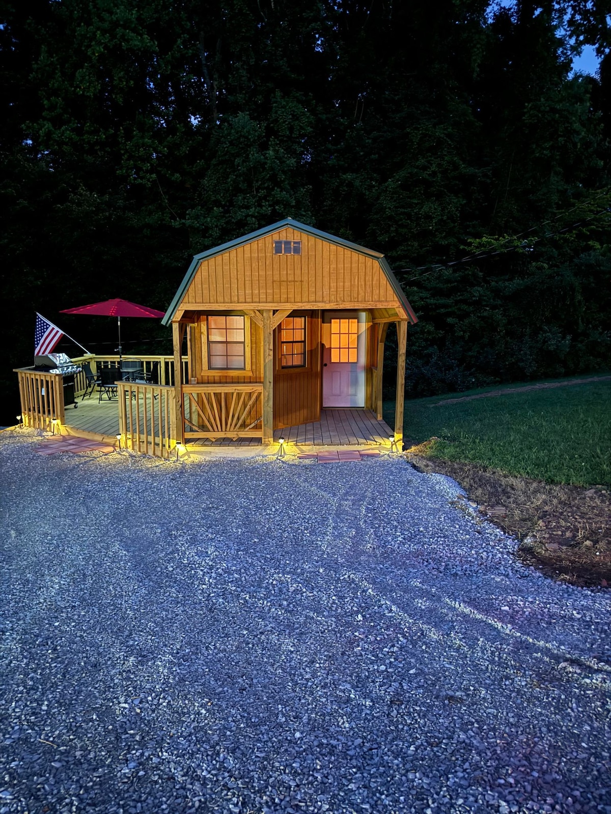 A charming tiny house, constructed in wood, is illuminated with soft lights, creating a welcoming ambiance. An umbrella provides shade over the outdoor dining area. A flag is displayed, adding a patriotic touch, while the gravel pathway leads to the front entrance.
