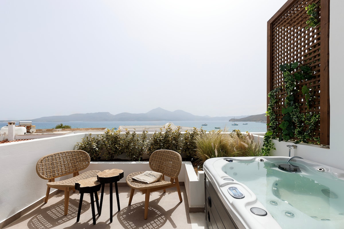 A private balcony features a relaxing jacuzzi and two wicker chairs with a small wooden table. Lush green plants cascade along a wooden lattice, enhancing the serene atmosphere. The expansive ocean views and distant mountains contribute to a peaceful outdoor setting.