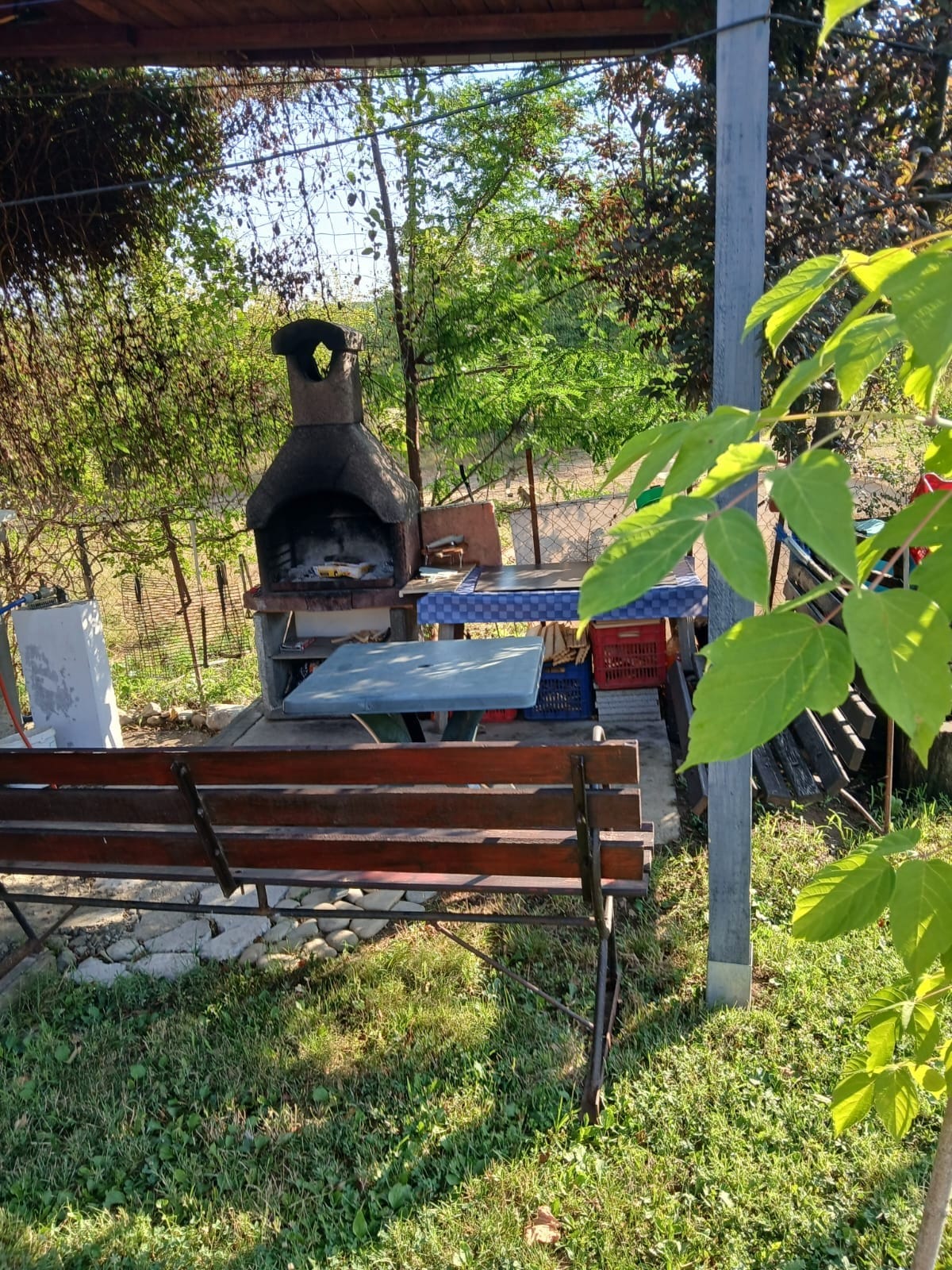 An outdoor cooking area is presented, featuring a traditional grill with a chimney. A wooden table is placed in front, accompanied by a bench. Lush greenery surrounds the space, contributing to a relaxed environment ideal for family gatherings.