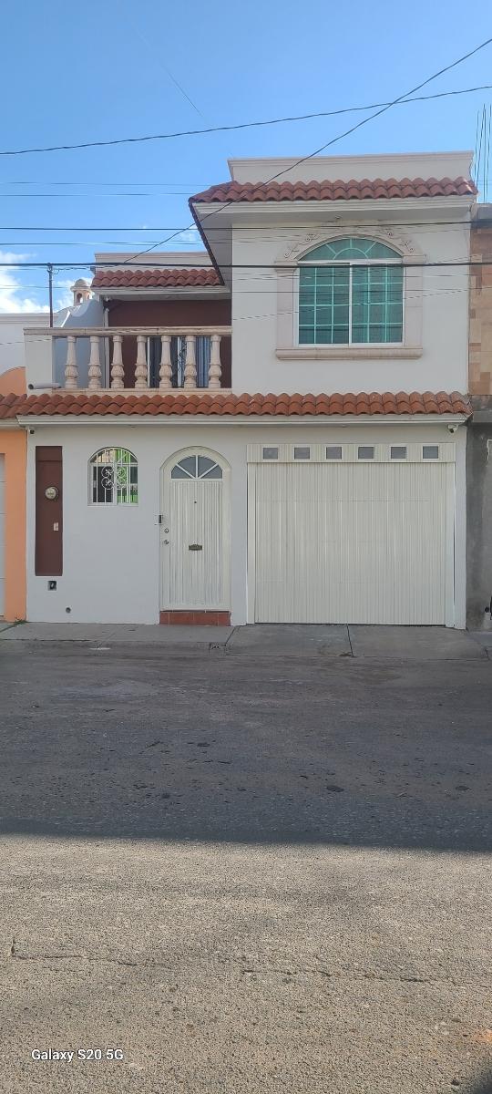 A two-story house exterior is presented, featuring a white facade with a tile roof. A decorative balcony extends over the entrance, which includes a round window. A garage door is visible, along with a smaller door adorned with a curved top.