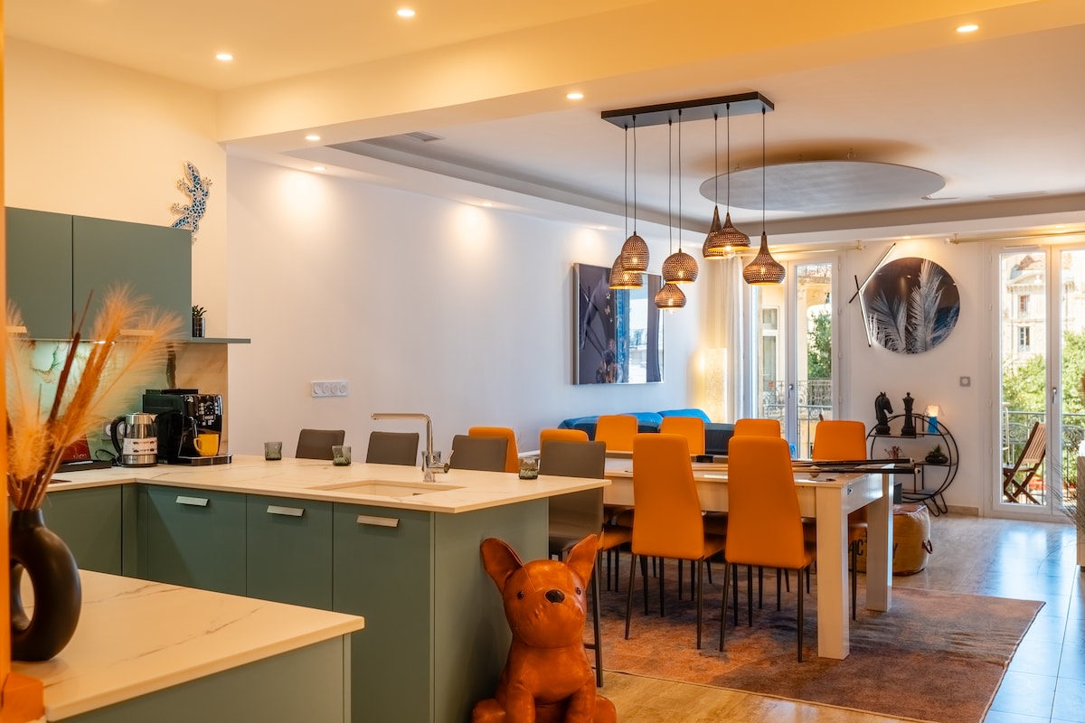 An open-plan living area is showcased, featuring a modern kitchen with a teal color scheme and a spacious dining table surrounded by brightly colored chairs. Artisan-style pendant lights hang above the dining area, while large windows invite natural light, revealing a glimpse of the outdoor balcony.